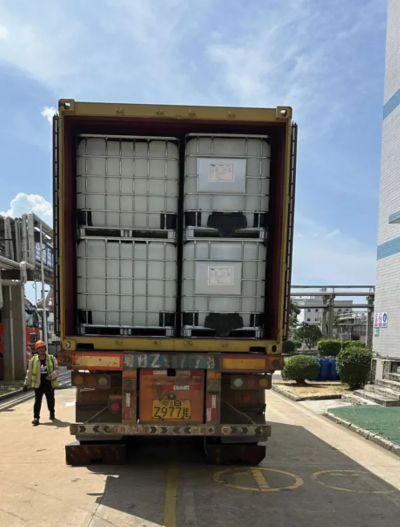 A shipping container loaded with large white industrial tanks is open at the back of a truck. Two workers in safety vests are nearby in an outdoor industrial area on a sunny day.
