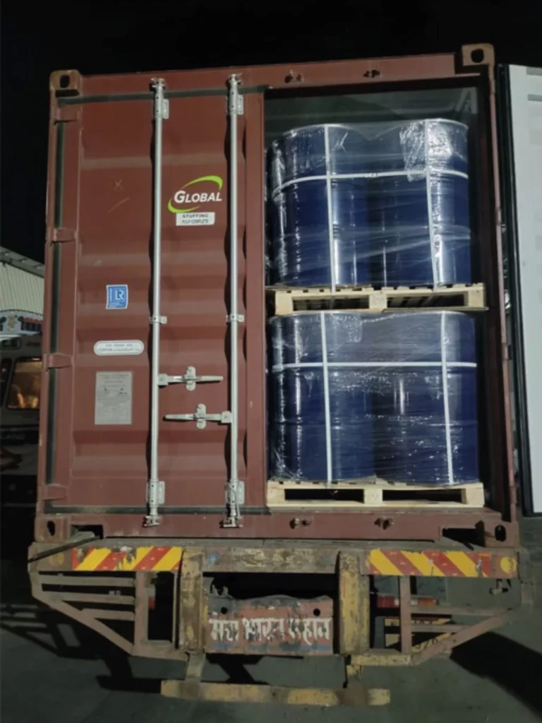 An open shipping container holds blue metal barrels stacked on wooden pallets and wrapped in plastic. The container is marked with a "Global" logo and is photographed at night.