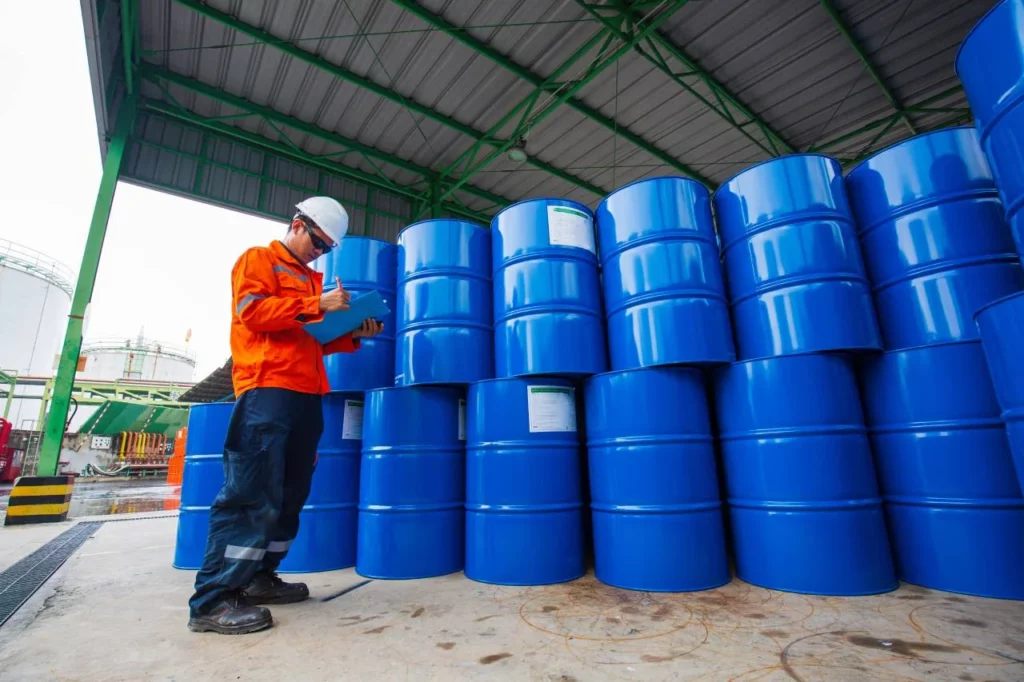 A worker in safety gear inspects and takes notes on stacked blue industrial barrels under a metal-roofed structure at an outdoor facility.