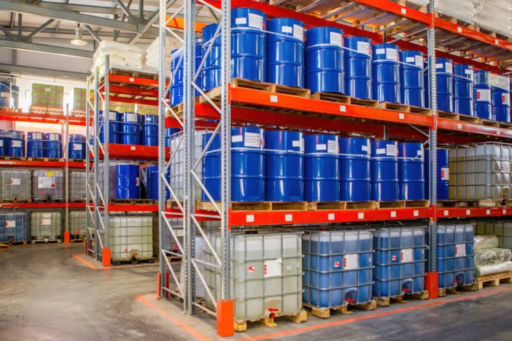Industrial warehouse with metal shelves stacked with large blue barrels and square plastic containers, storing various chemicals. The area is organized and well-lit, with safety labels visible on containers.