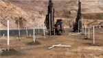 Two large drilling machines operate on a dirt construction site with PVC pipes sticking out of the ground, possibly for soil sampling or marking drilling locations. Rocky, barren hills rise in the background.